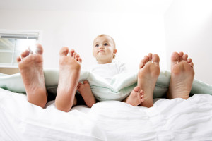 This is a photo of a mom and dad on either side of their one year old baby in bed together. There is a lot of space for copy above.